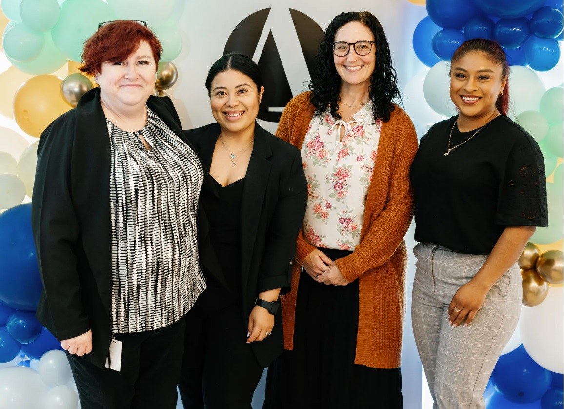 Four women in casual office attire smiling at camera standing in front of Alleviate Logo.